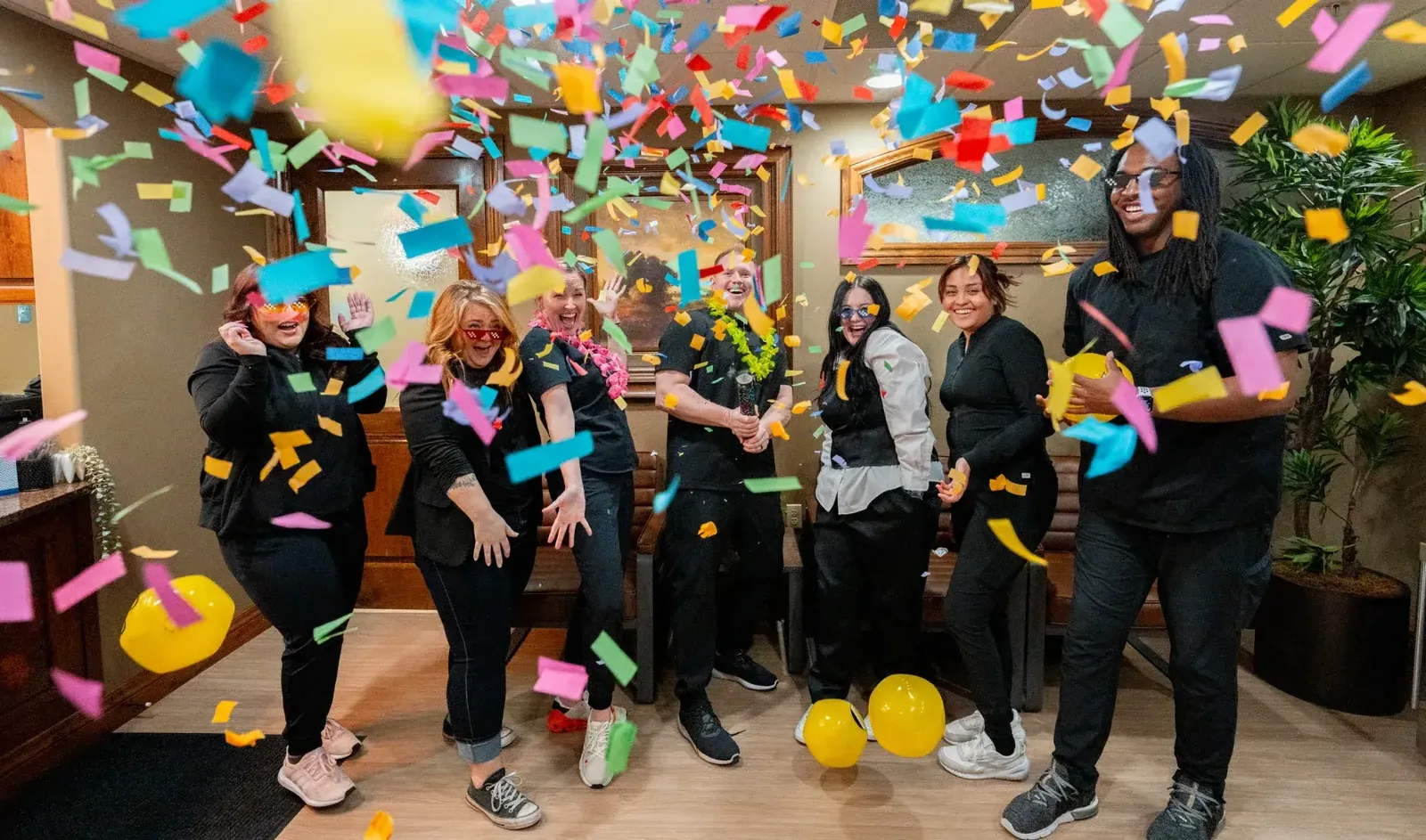 Dental staff members celebrating with balloons under falling colorful confetti in an office lobby.
